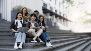 Group of cheerful Asian college students sitting on stairs, showing fists, celebrating triumph